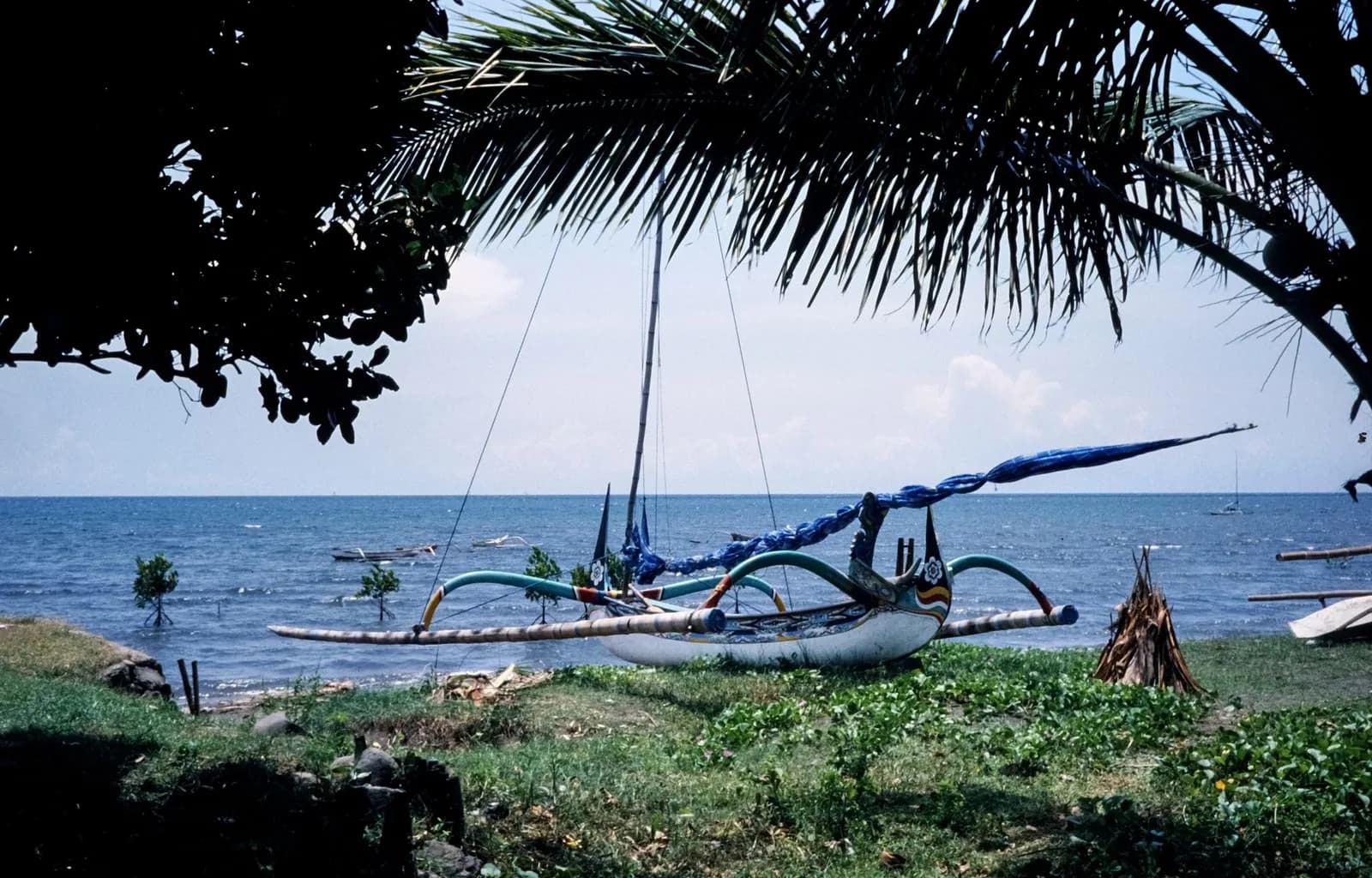 Jukung traditionnel balinais à balancier peint, posé sur l'herbe face à la mer calme de la côte est de Bali