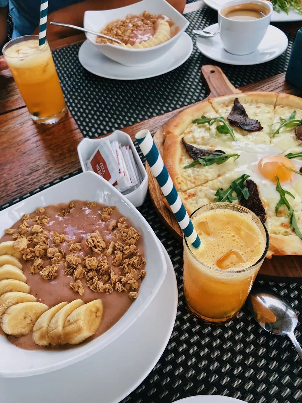 Table de brunch dans un café de Bali avec smoothie bowls à la banane et au granola, jus pressés et pizza aux œufs