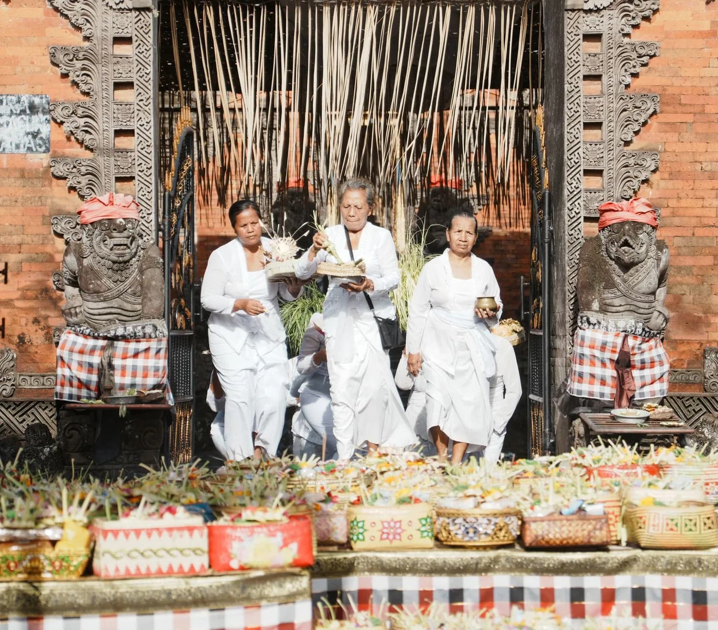 Femmes balinaises vêtues de blanc portant des offrandes devant la porte d'un temple, entre deux gardiens de pierre