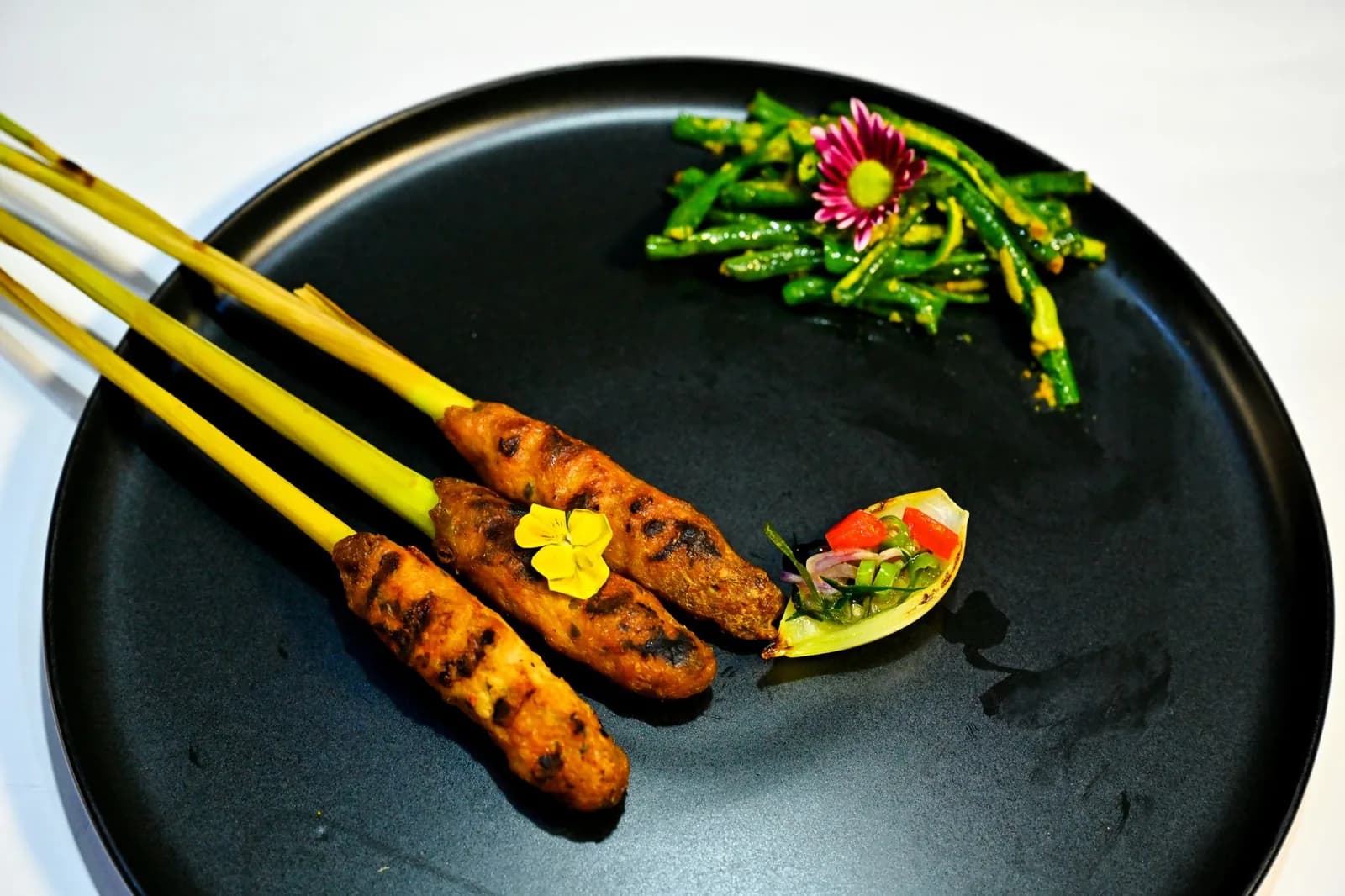 Trois brochettes de sate lilit balinais enroulées autour de bâtons de citronnelle, servies avec du sambal matah et des haricots longs en urap
