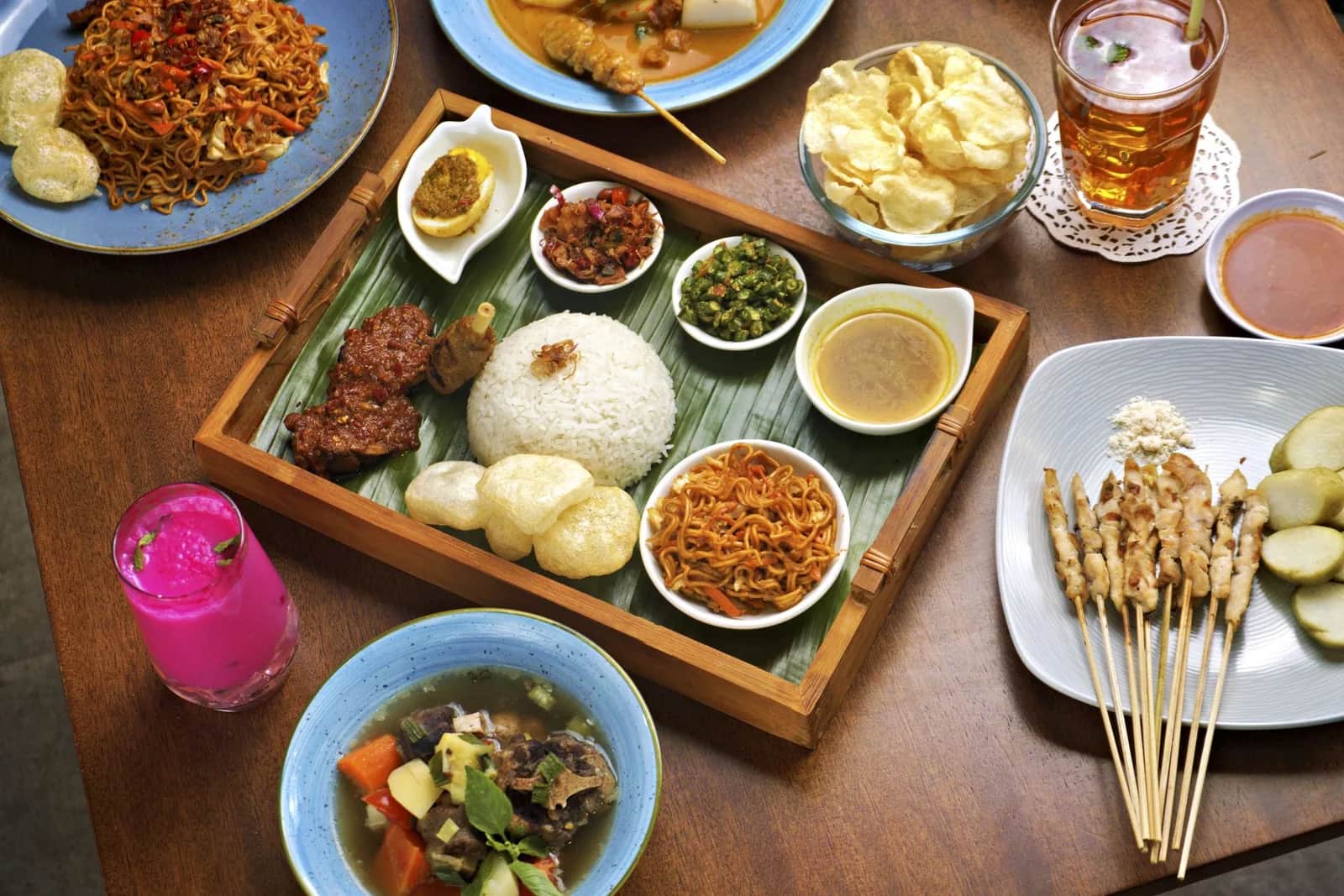 Table couverte de plats indonésiens à Bali : plateau de nasi campur sur feuille de bananier, brochettes de satay, mie goreng, kerupuk, soupe et jus de fruit frais