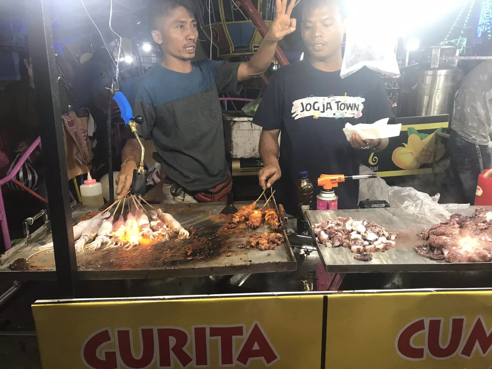 Deux vendeurs grillent des brochettes et des fruits de mer sur la plaque d'un stand du marché de nuit d'Ubud, à Bali