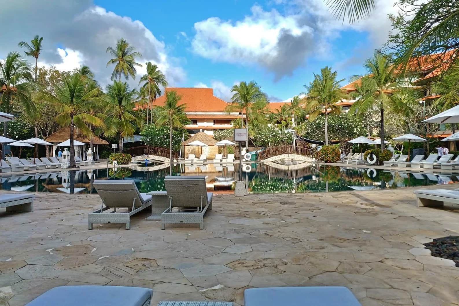 Piscine d'un grand hôtel balinais bordée de palmiers, transats et toits de tuiles orange