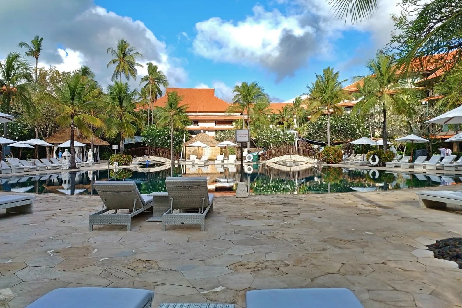 Piscine d'un grand hôtel balinais bordée de palmiers, transats et toits de tuiles orange