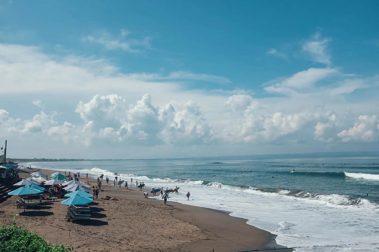 Plage de sable sombre sur la côte de Canggu, avec parasols turquoise, surfeurs et vagues sous un ciel nuageux