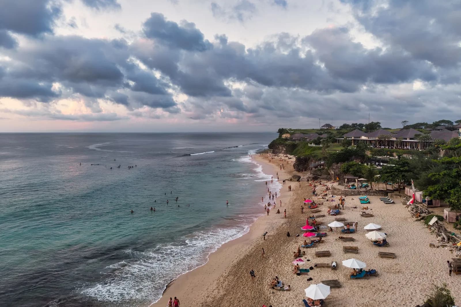 Plage de sable au pied de la falaise d'Uluwatu, avec baigneurs, parasols et un resort sur le promontoire, au crépuscule