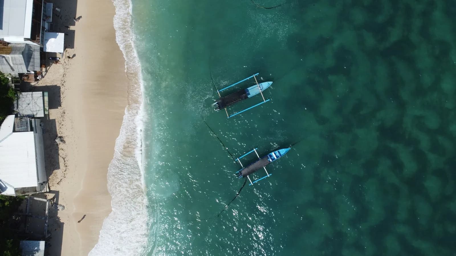 Vue aérienne d'une baie balinaise à l'eau claire et calme, deux bateaux de pêche à balancier ancrés devant les warungs de la plage
