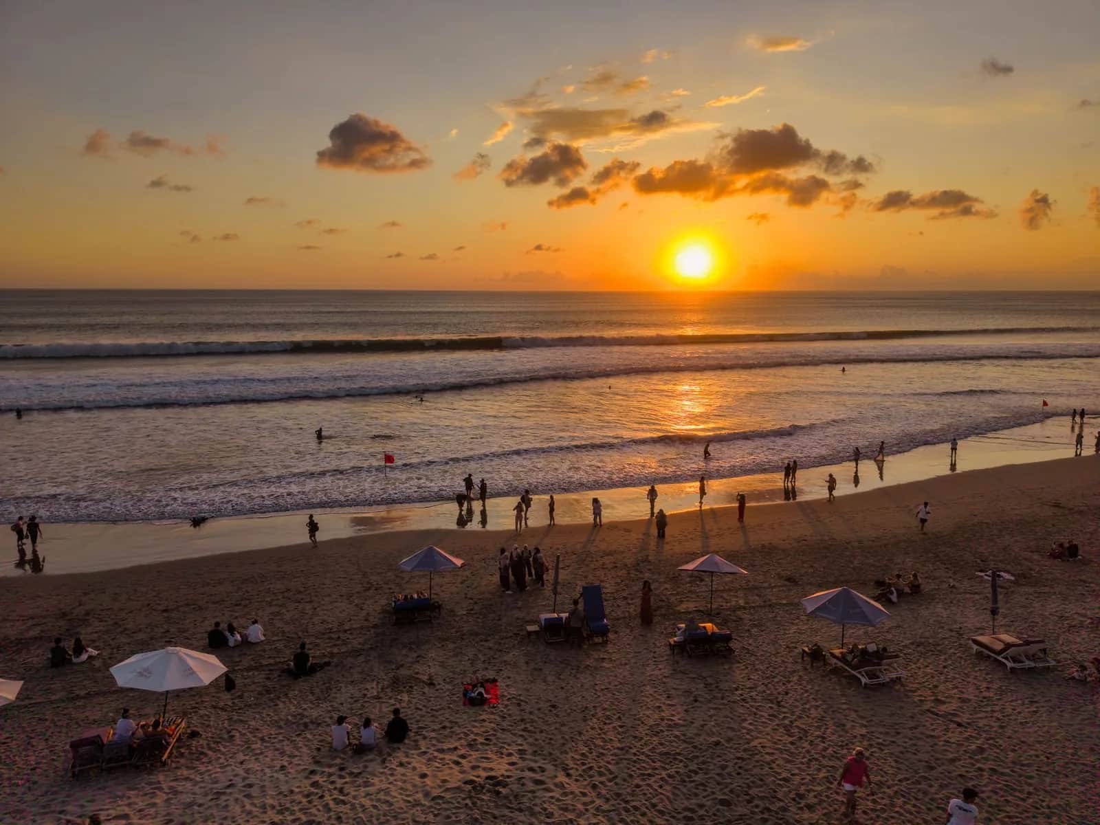 Coucher de soleil sur la plage de Kuta à Bali, parasols et transats sur le sable, baigneurs dans les rouleaux et drapeau rouge planté au bord de l'eau