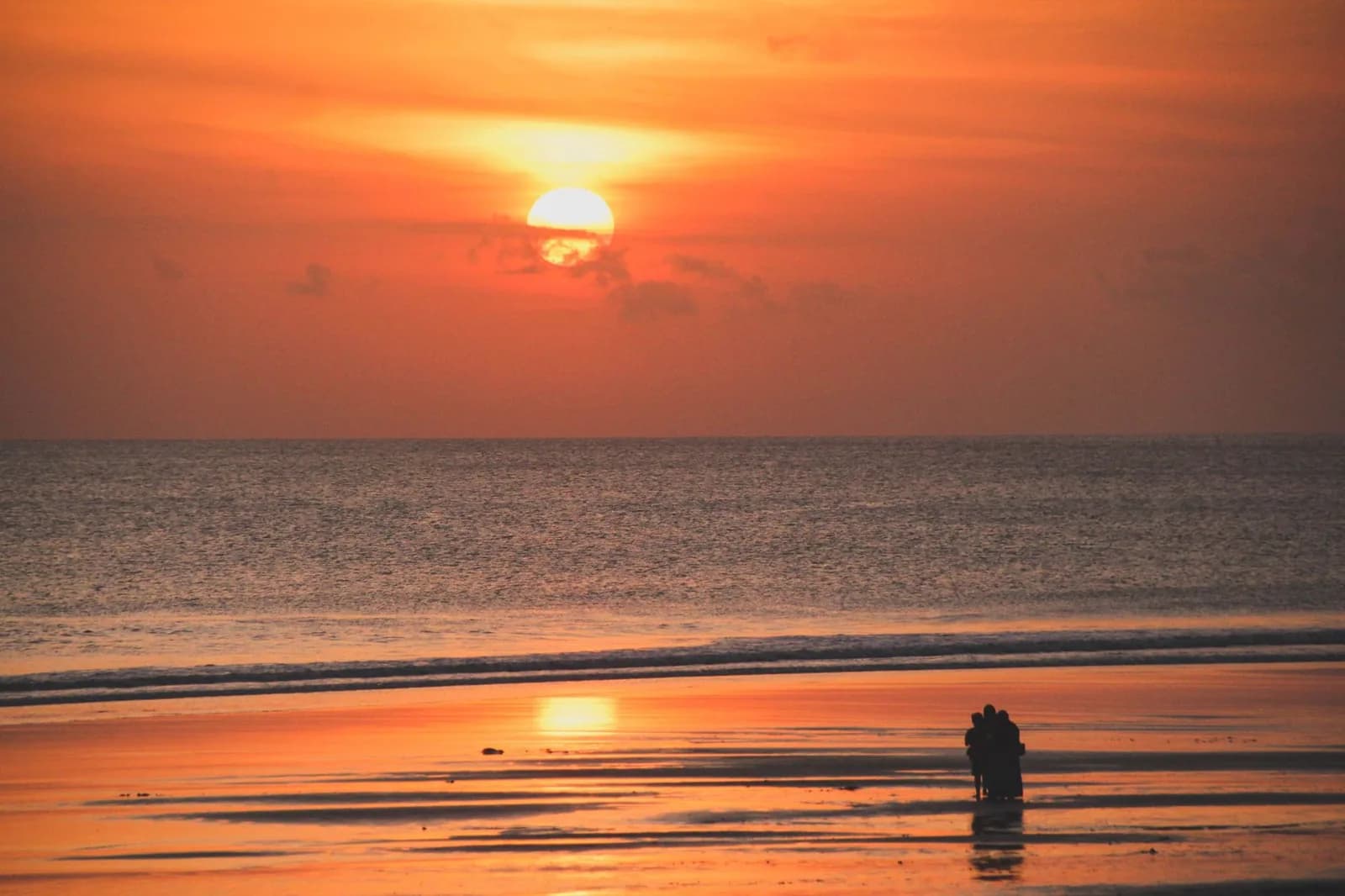 Soleil couchant sur la plage de Kuta à Bali à marée basse, silhouettes d'un groupe sur le sable mouillé et reflets orange jusqu'à l'horizon