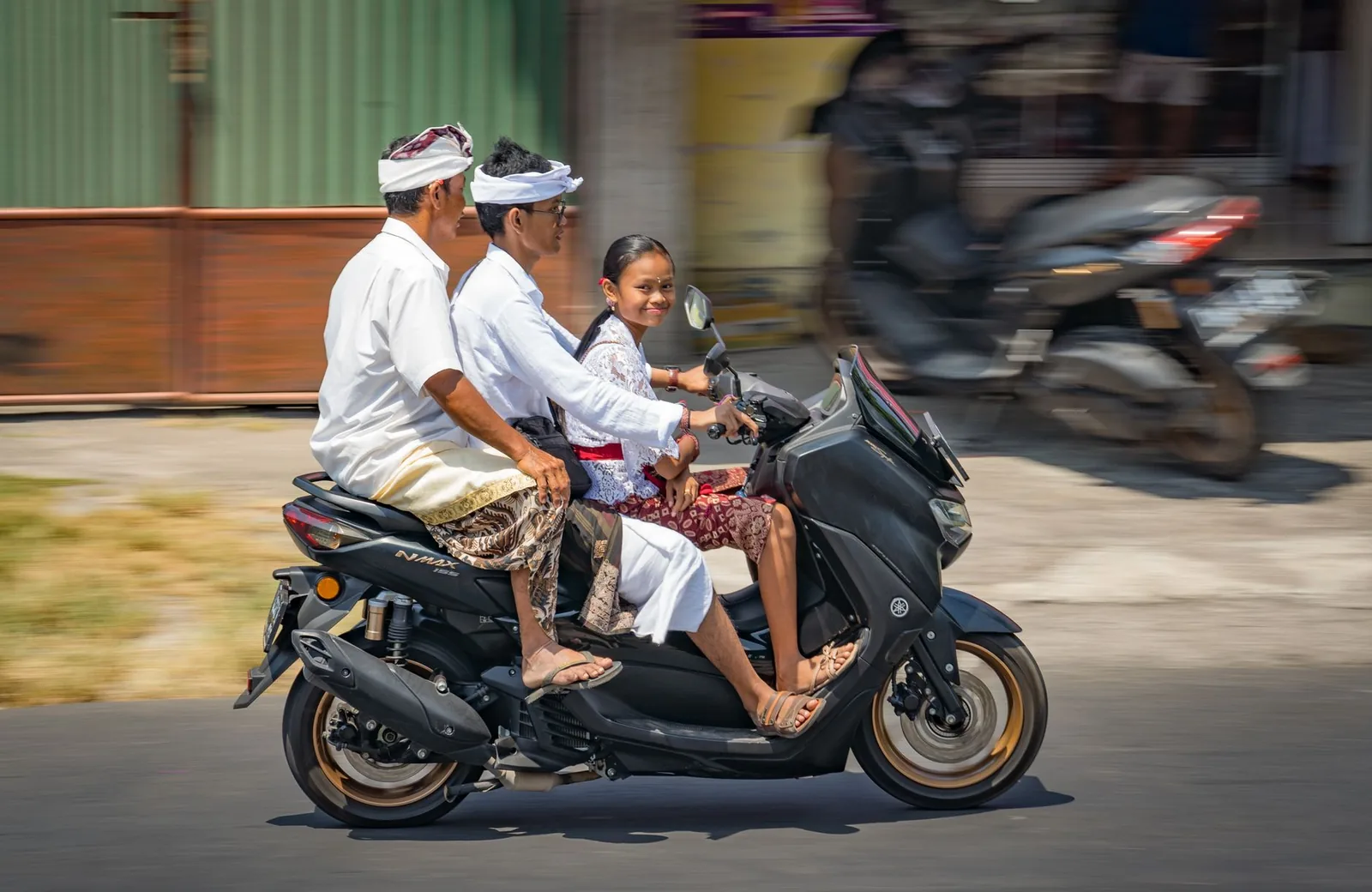 Famille balinaise en tenue de cérémonie roulant à trois sur un Yamaha NMAX 155 dans une rue de Bali