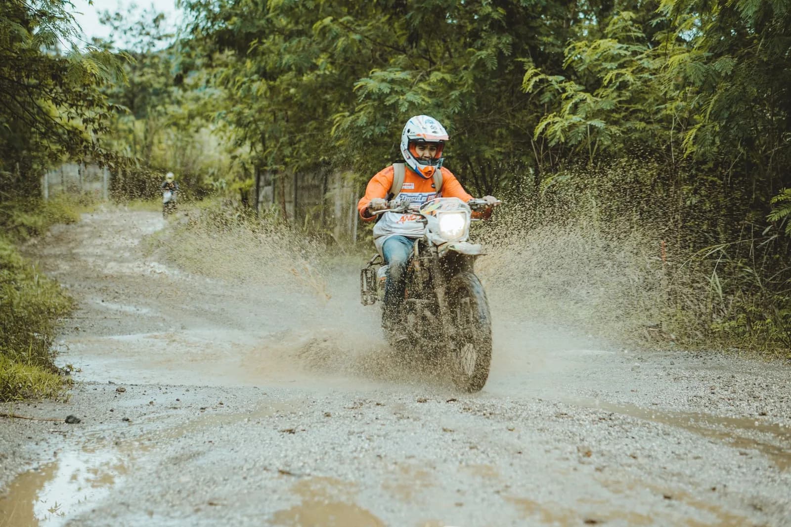 Pilote d'enduro lançant une gerbe de boue sur une piste de terre bordée de végétation tropicale dense