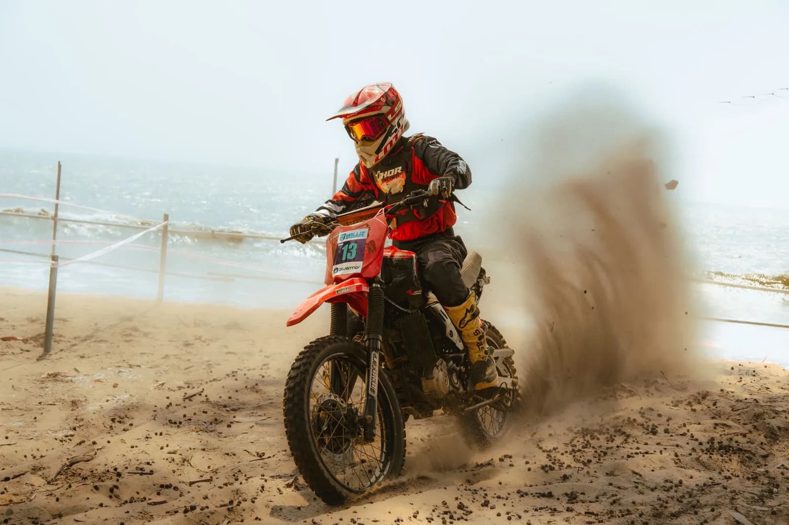Pilote de moto tout-terrain en tenue rouge dérapant sur le sable d'une plage, la mer et l'écume derrière lui