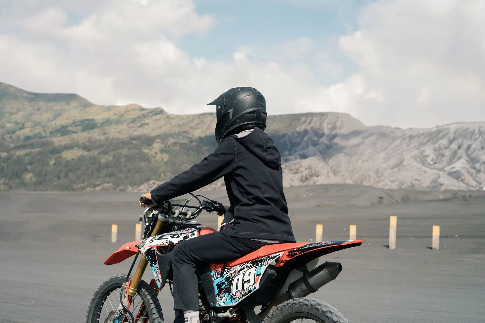 Pilote arrêté sur sa moto tout-terrain au milieu d'une vaste plaine de cendre volcanique noire, face aux parois d'une caldeira