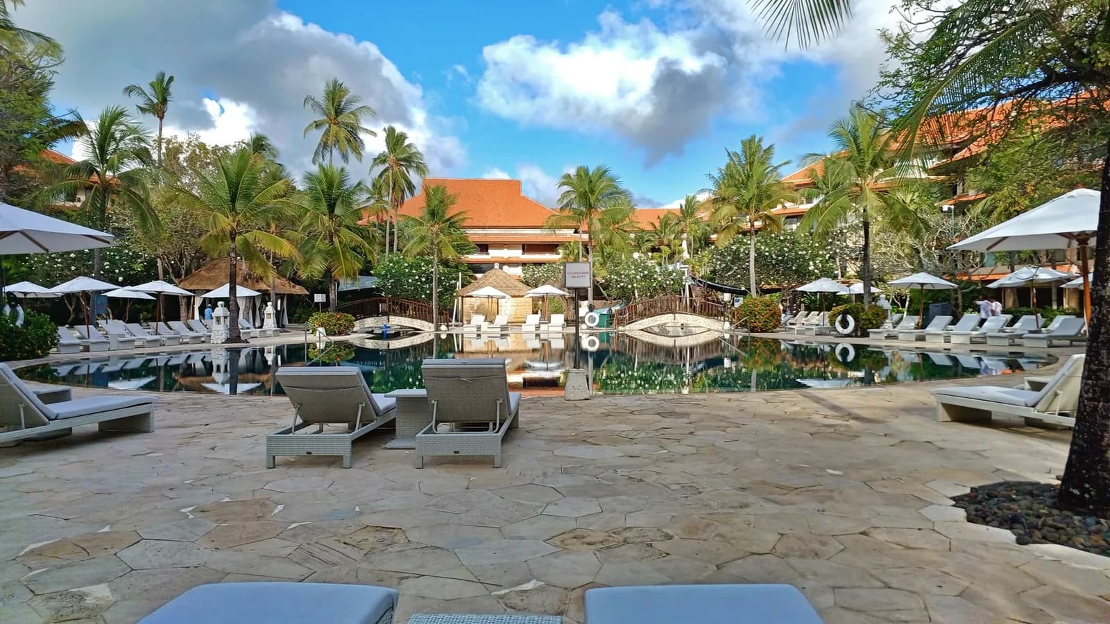 Piscine et transats d'un grand resort de Nusa Dua à Bali, toits de tuiles orange, cocotiers et frangipaniers autour du bassin