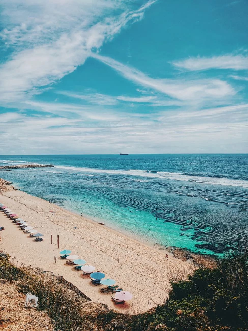 Plage de sable blanc en contrebas d'une falaise du Bukit, parasols colorés et récif visible sous l'eau turquoise au sud de Bali