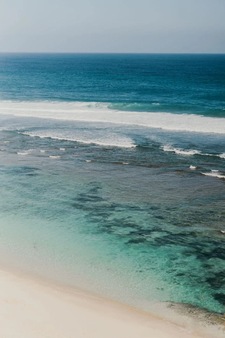 Récif frangeant de Nusa Dua vu du dessus : la houle se brise au large sur la barrière, l'eau du lagon reste claire et peu profonde près du sable blanc