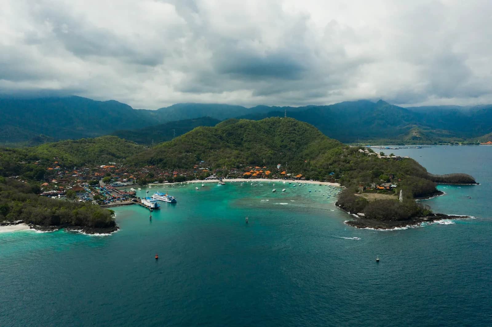 Vue aérienne de la baie de Padang Bai à Bali, avec le terminal ferry à gauche, le village et la plage au fond de la baie et le cap est couvert de forêt