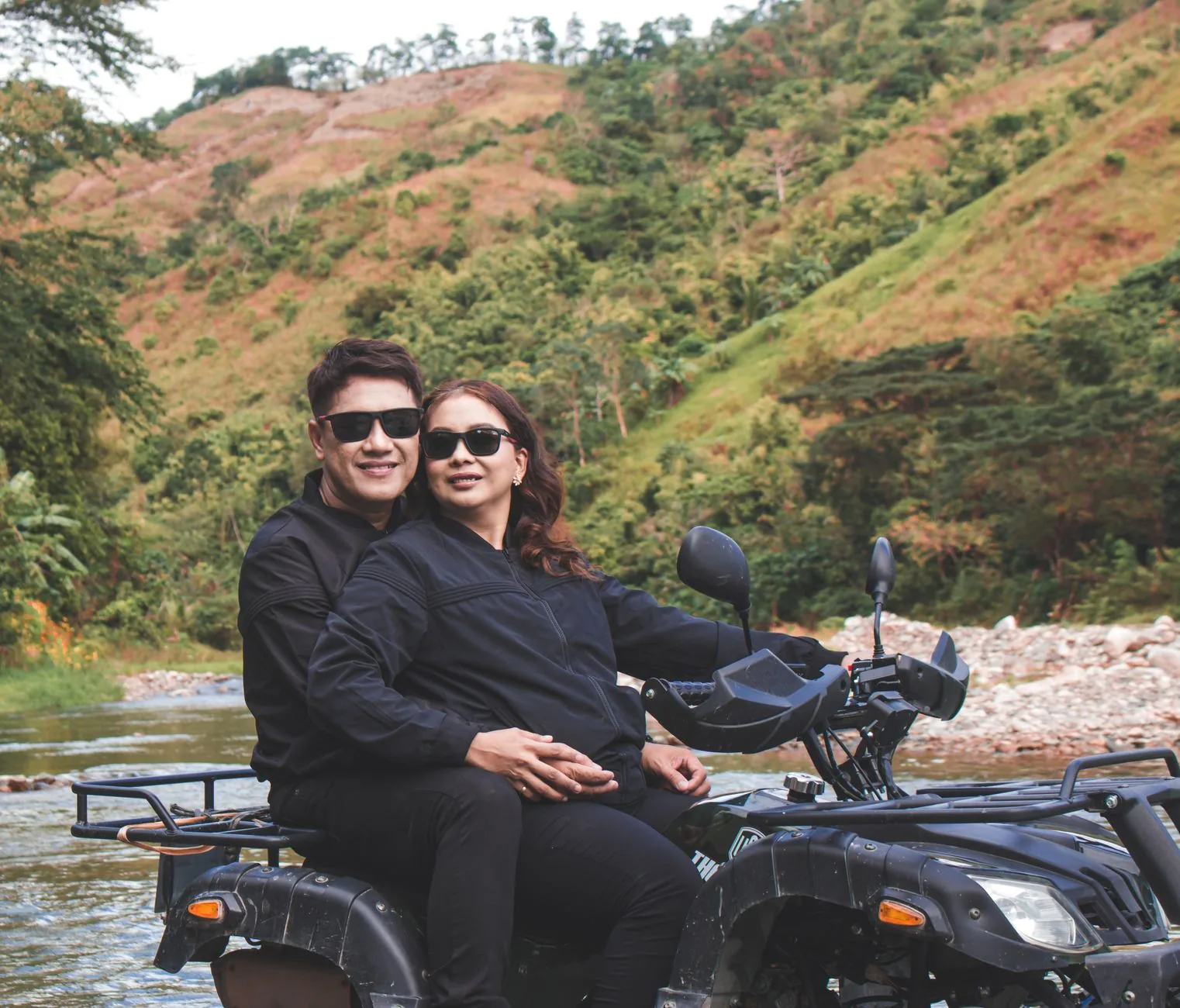 Couple assis sur un quad tout-terrain au bord d'une rivière, devant des collines couvertes de végétation tropicale