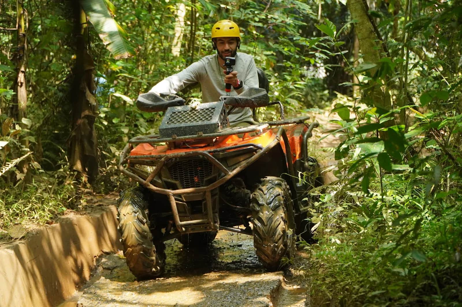 Pilote en casque jaune au guidon d'un quad orange franchissant un passage humide sur un sentier de jungle tropicale, comme lors d'une sortie ATV près d'Ubud à Bali