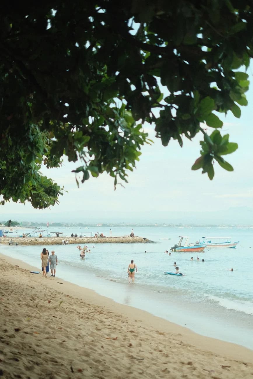Baigneurs dans l'eau calme du lagon de Sanur à marée haute, jukung au mouillage et jetée de pierre au loin