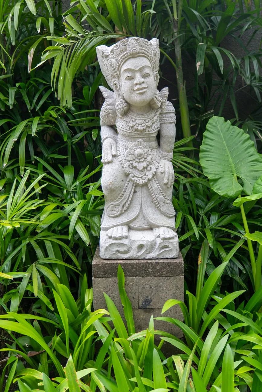 Statue de pierre balinaise sculptée posée sur un socle au milieu d'une végétation tropicale dense, comme au Big Garden Corner de Sanur