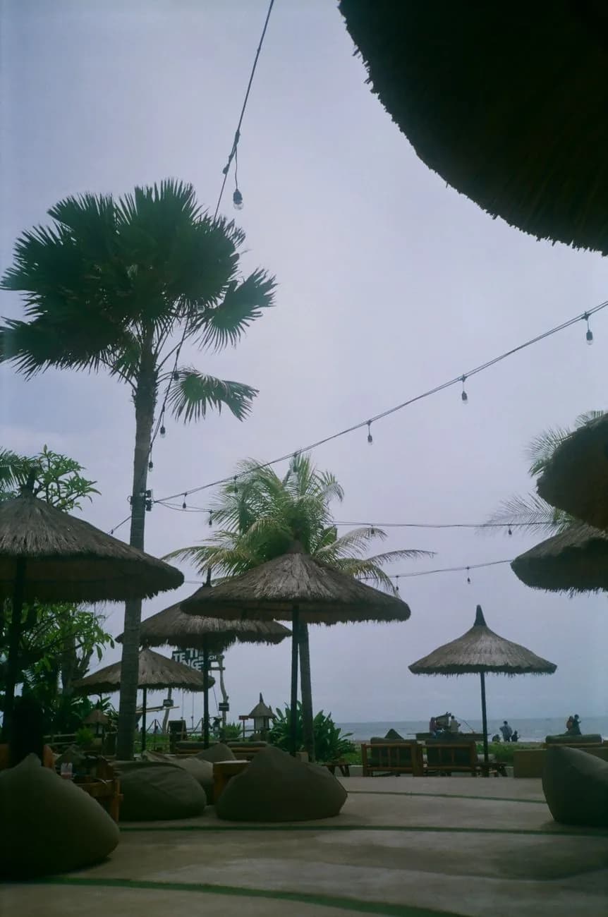 Beach club en bord de mer à Seminyak, parasols de paille, poufs posés sur le sol et guirlandes lumineuses face à l'océan