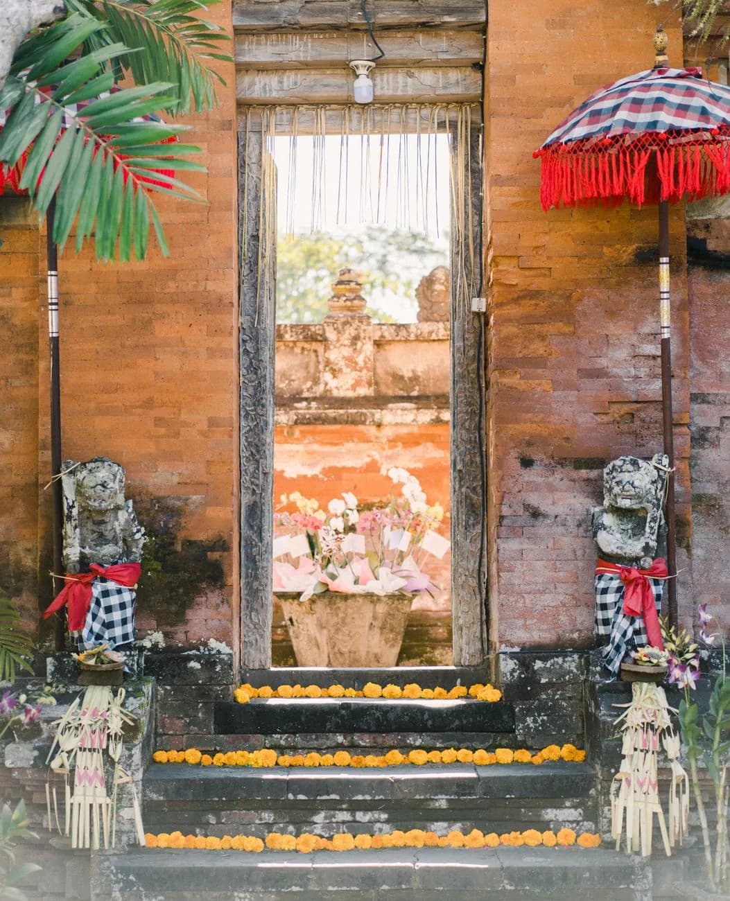 Porte de temple balinais en brique rouge décorée d'offrandes de fleurs, d'une ombrelle cérémonielle et de gardiens de pierre, comme au temple de Petitenget à Seminyak
