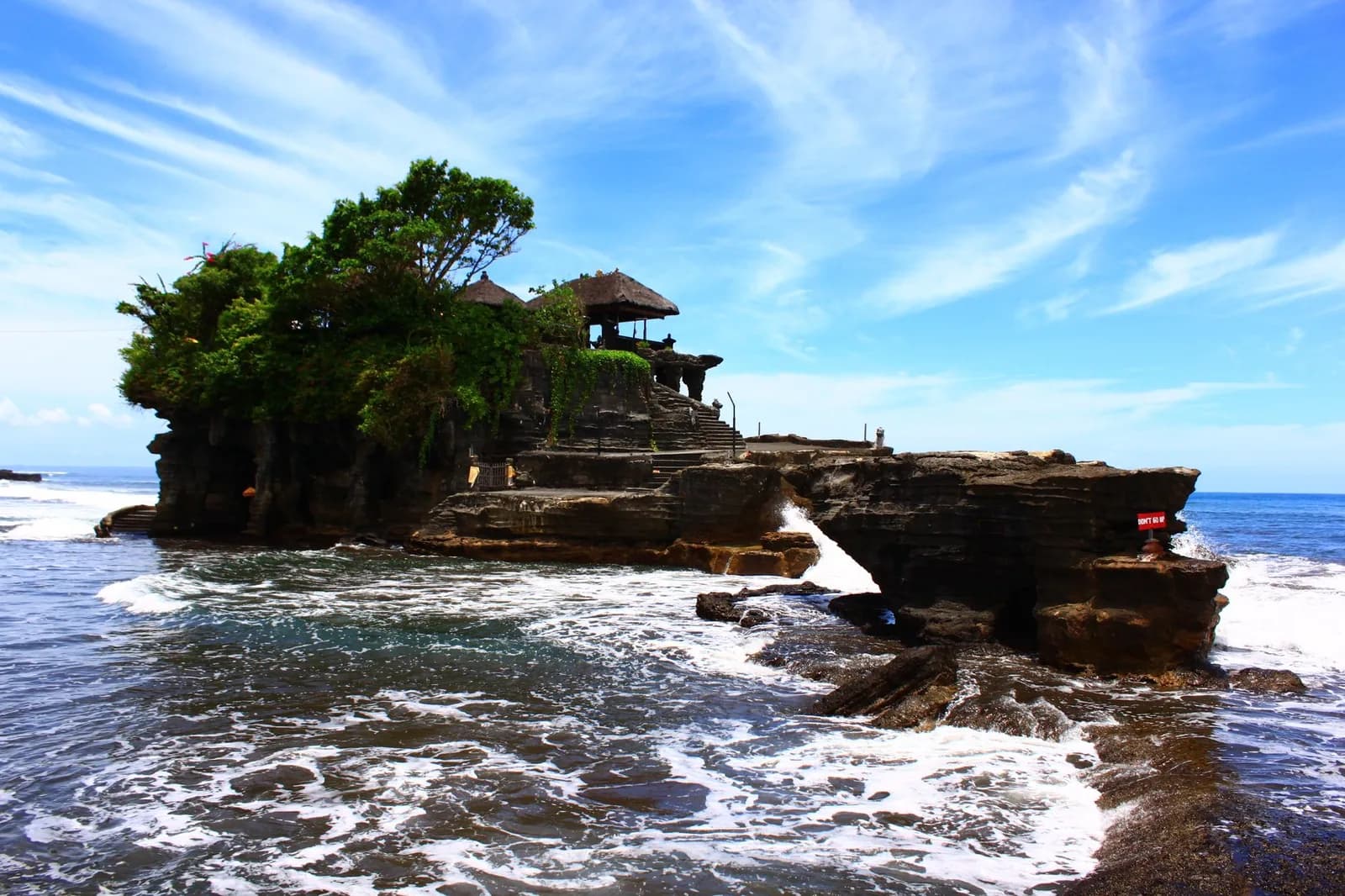 Le rocher du temple de Tanah Lot en pleine journée sous un ciel bleu, les vagues autour de sa base, Bali